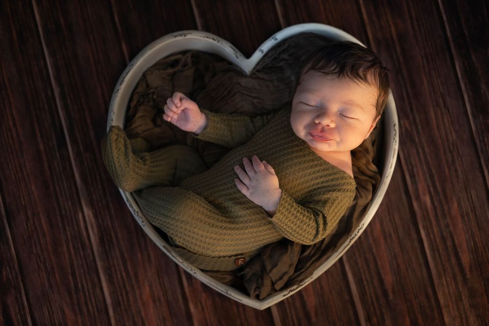 Baby sleeping peacefully in heart-shaped frame against dark wooden background.
