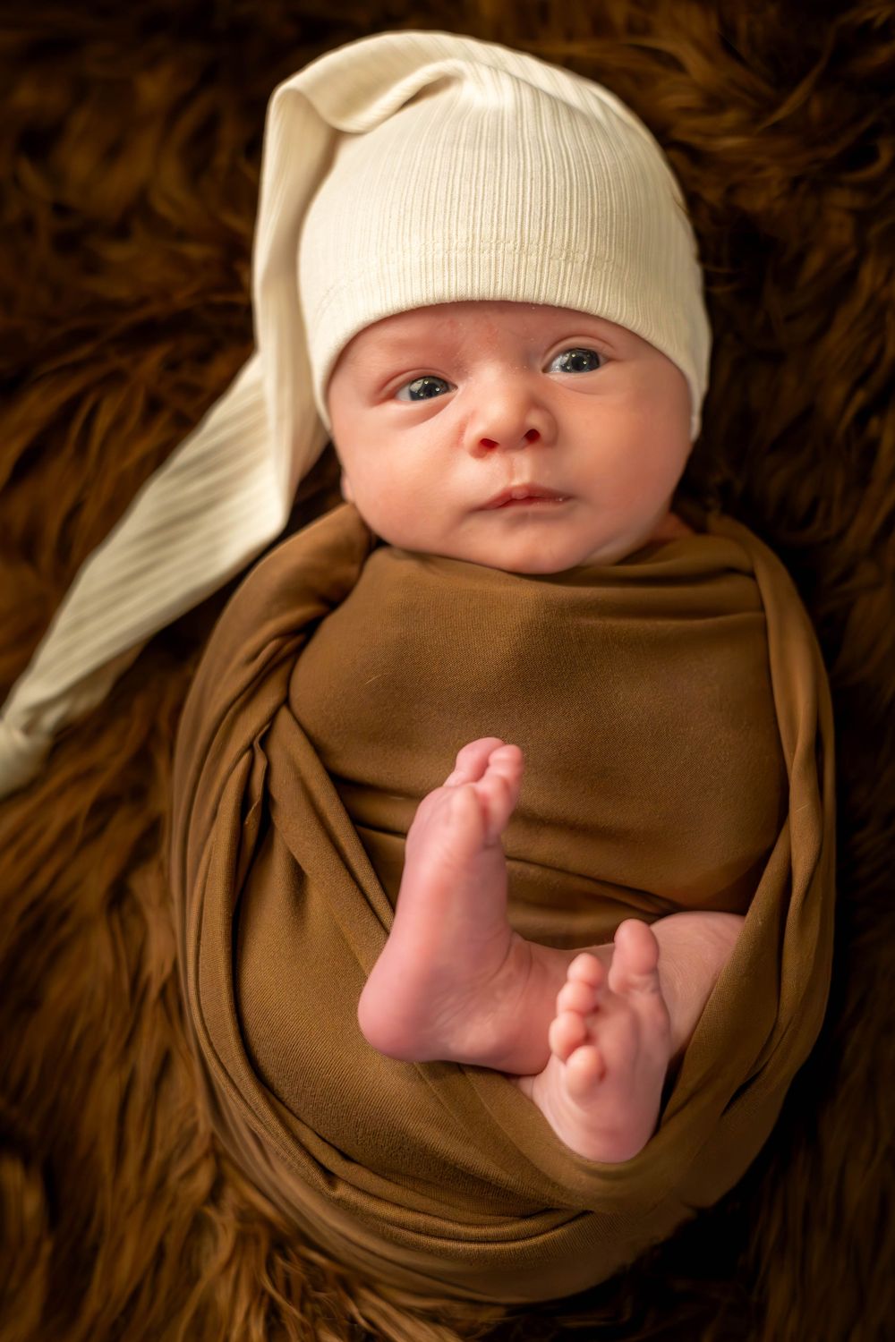 A series of photos showing a swaddled baby in a brown wrap with tiny feet peeking out, wearing a cream colored hat.