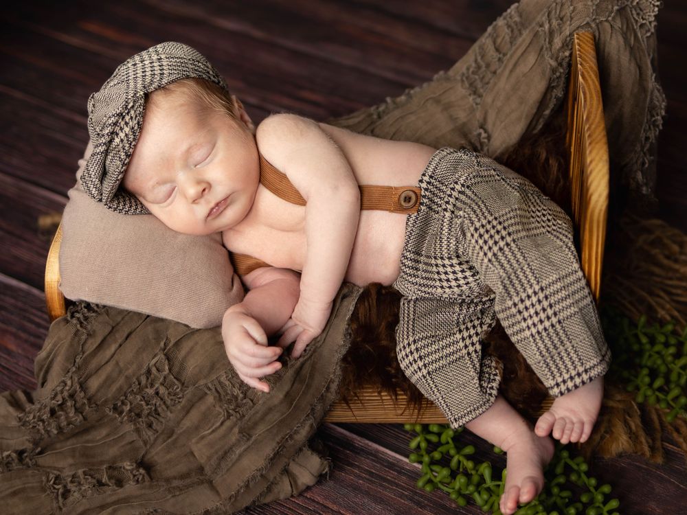 A sleeping baby lies on a wooden deck with a plaid blanket and greenery in the background.