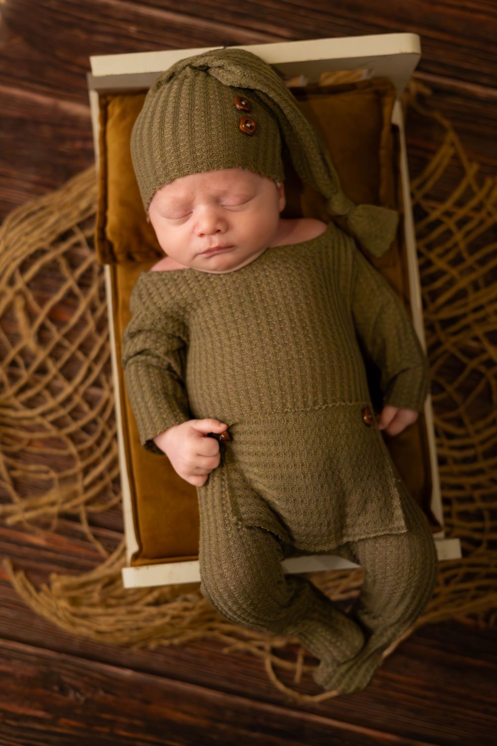 A sleeping baby wearing a knit olive green outfit and hat lies peacefully on a wooden surface with woven decor.