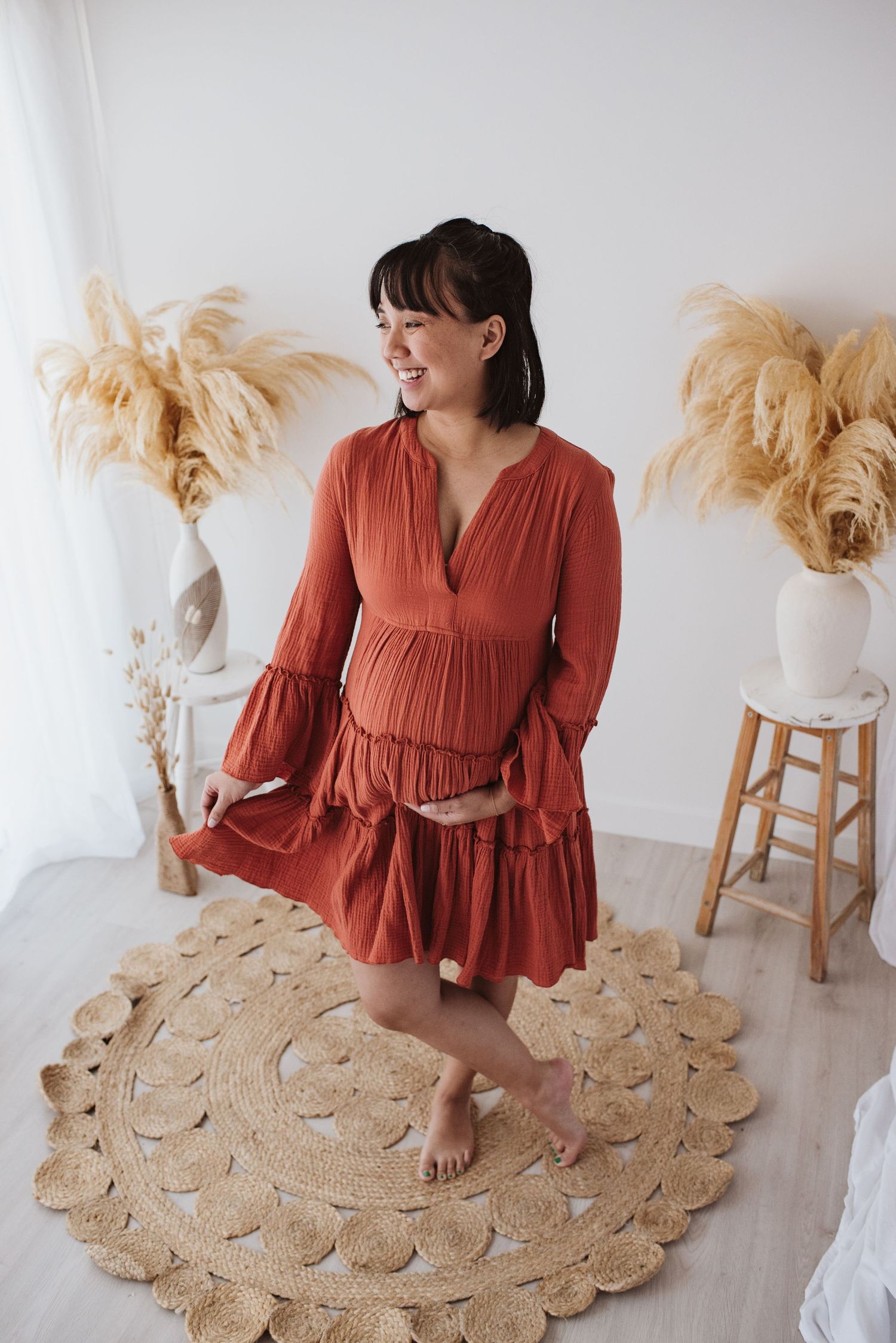 A rustic maternity photo in a coral-colored dress with bell sleeves on a woven jute rug.