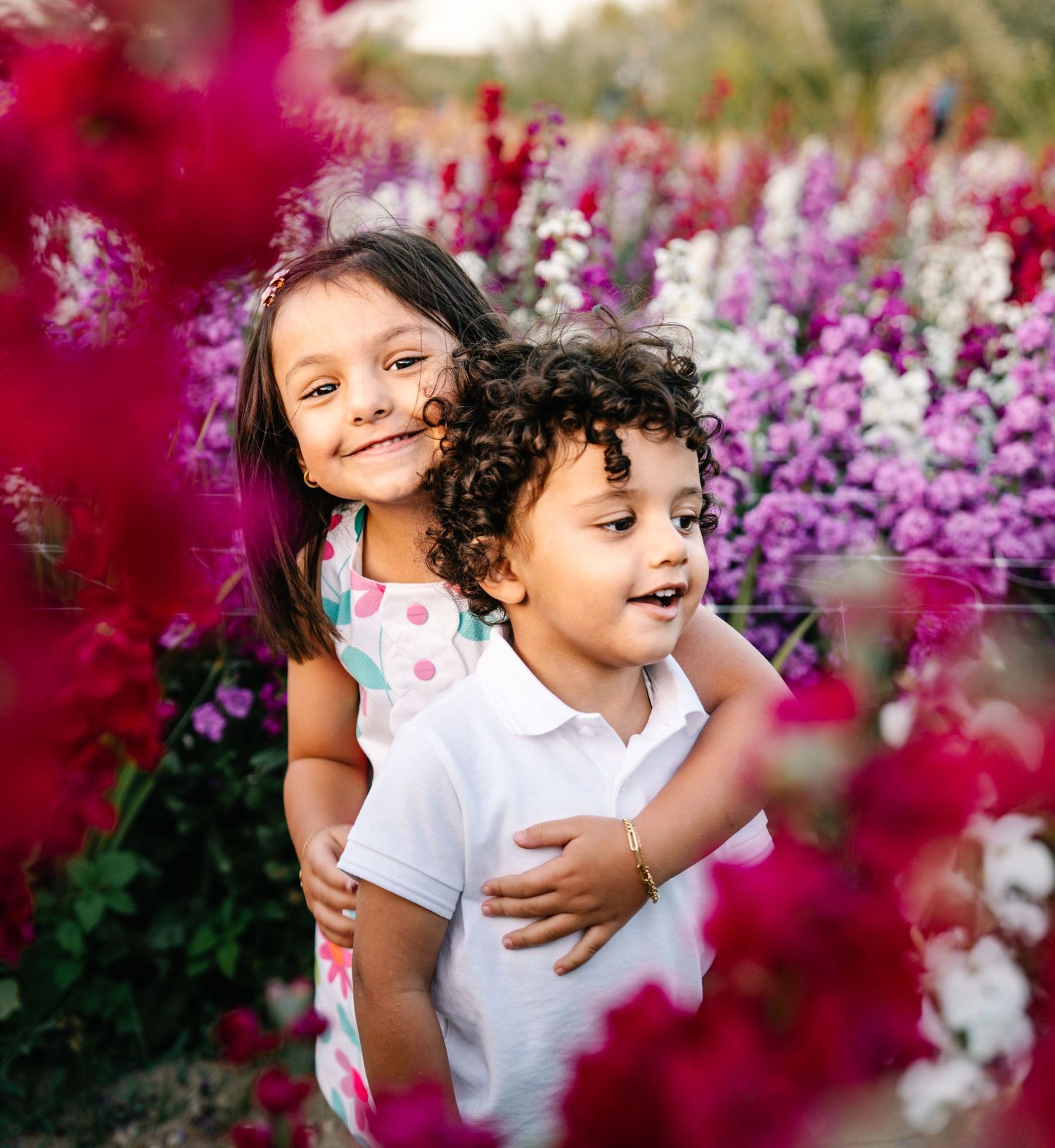 Two children embrace and smile among vibrant purple and red flowers.