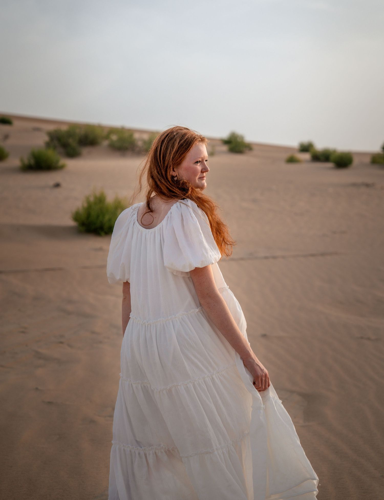 Woman in white flowy dress walks through desert dunes at golden hour.
