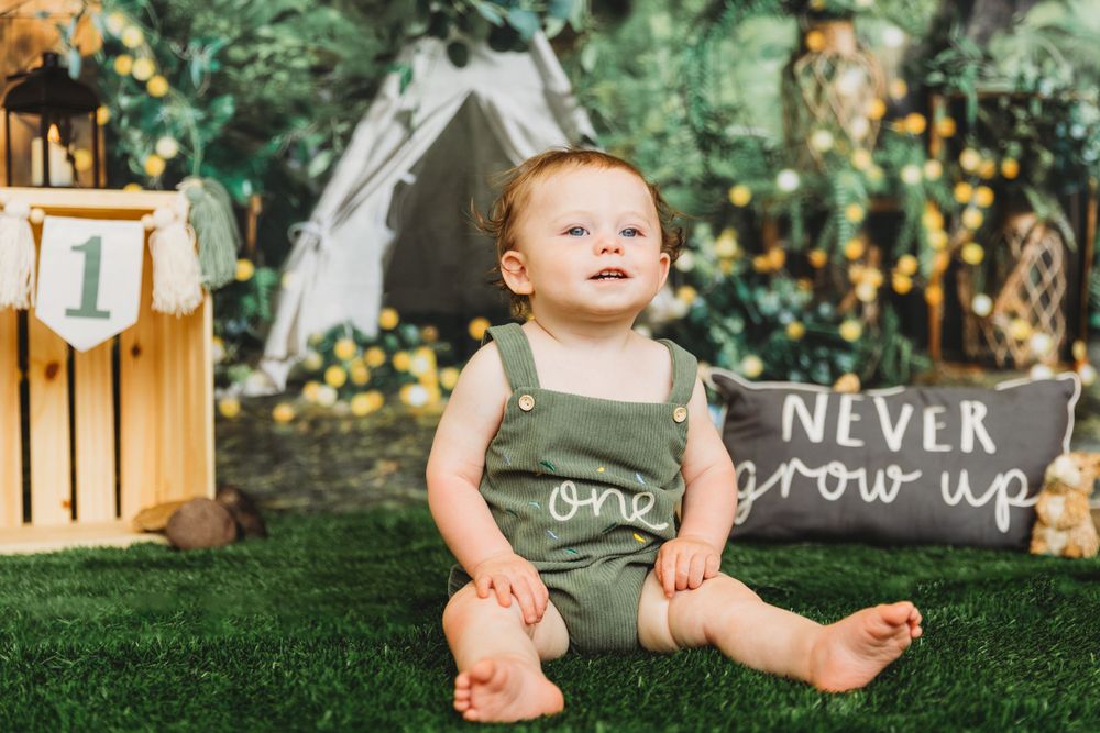 Baby in olive green romper sitting on grass with rustic party decorations.
