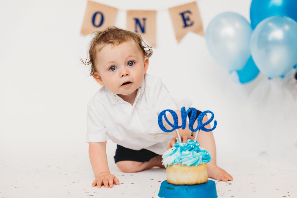 Baby in white shirt reaching for blue birthday cake with 'one' topper.
