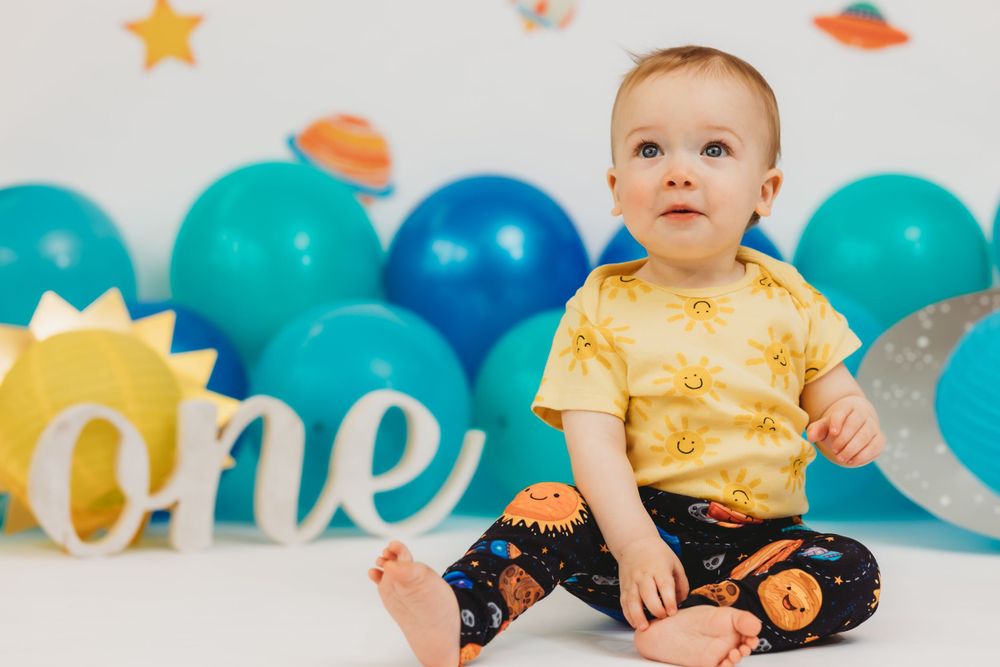 Baby in yellow shirt sitting against space-themed backdrop with blue balloons and 'one' text.