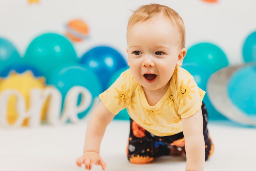Smiling baby in yellow shirt crawling on floor with blue balloons and space decorations.