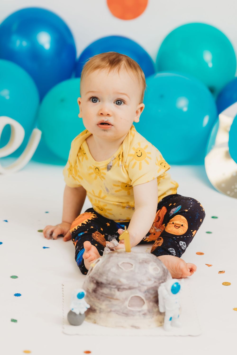Baby in yellow shirt sitting on floor with blue balloons and space-themed first birthday decorations.