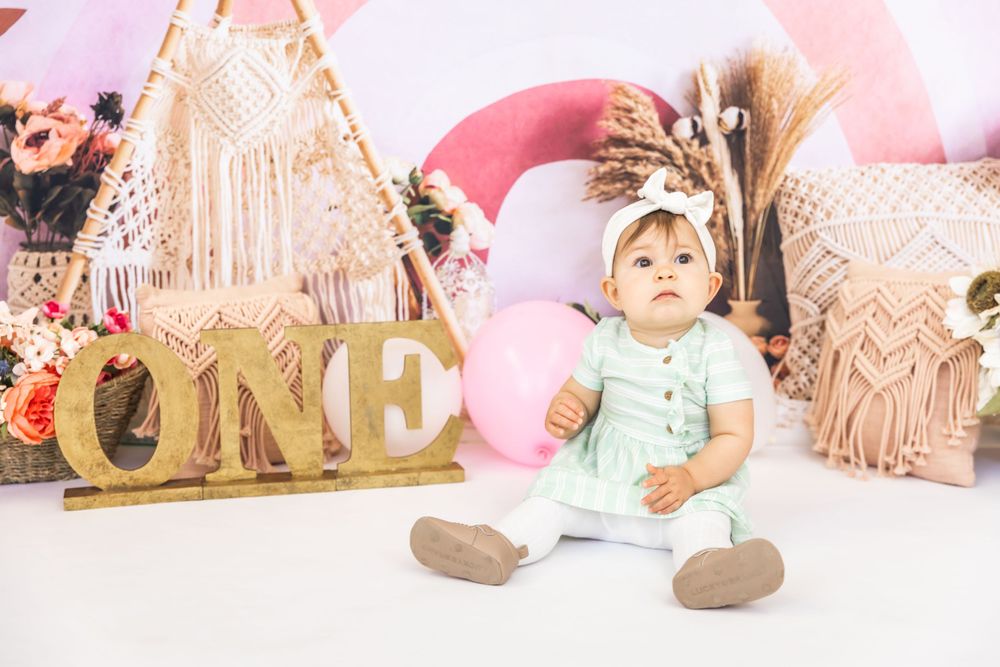 Baby in mint green outfit sits on floor with gold 'ONE' letters and boho decorations in pink setting.