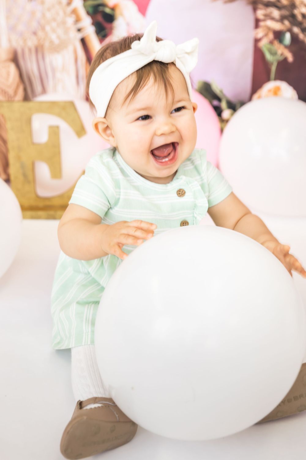 Baby in mint dress plays with large white balloon against pink backdrop with macrame decorations.