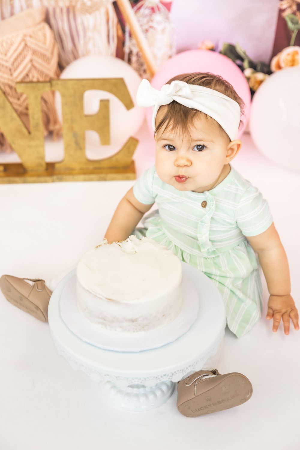 Baby in mint green outfit reaching for white birthday cake on pedestal.