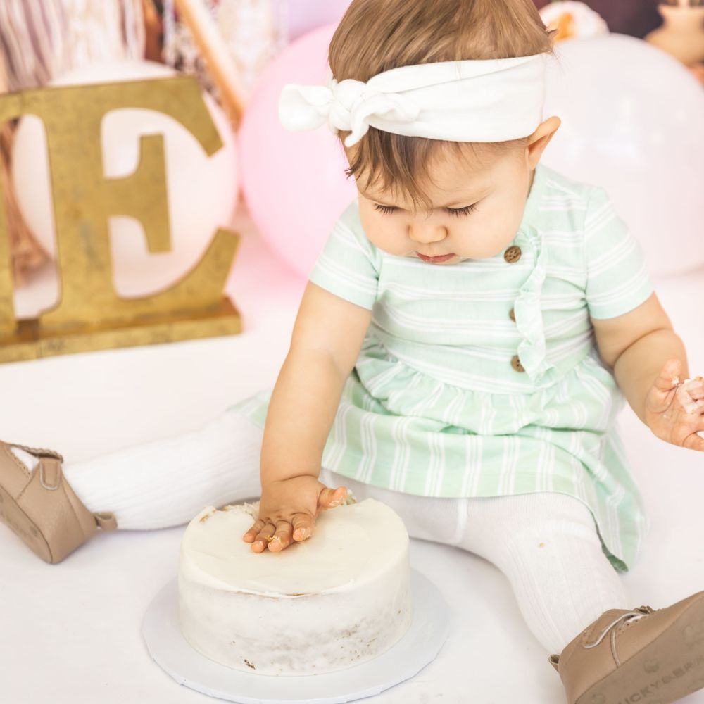 Baby in mint dress touching white cake during first birthday photoshoot.