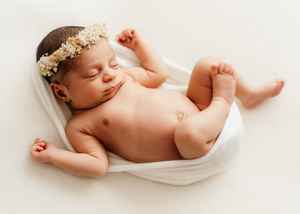 Peaceful sleeping newborn baby wearing delicate floral crown wrapped in soft white fabric on light background. Newborn Photoshoot.