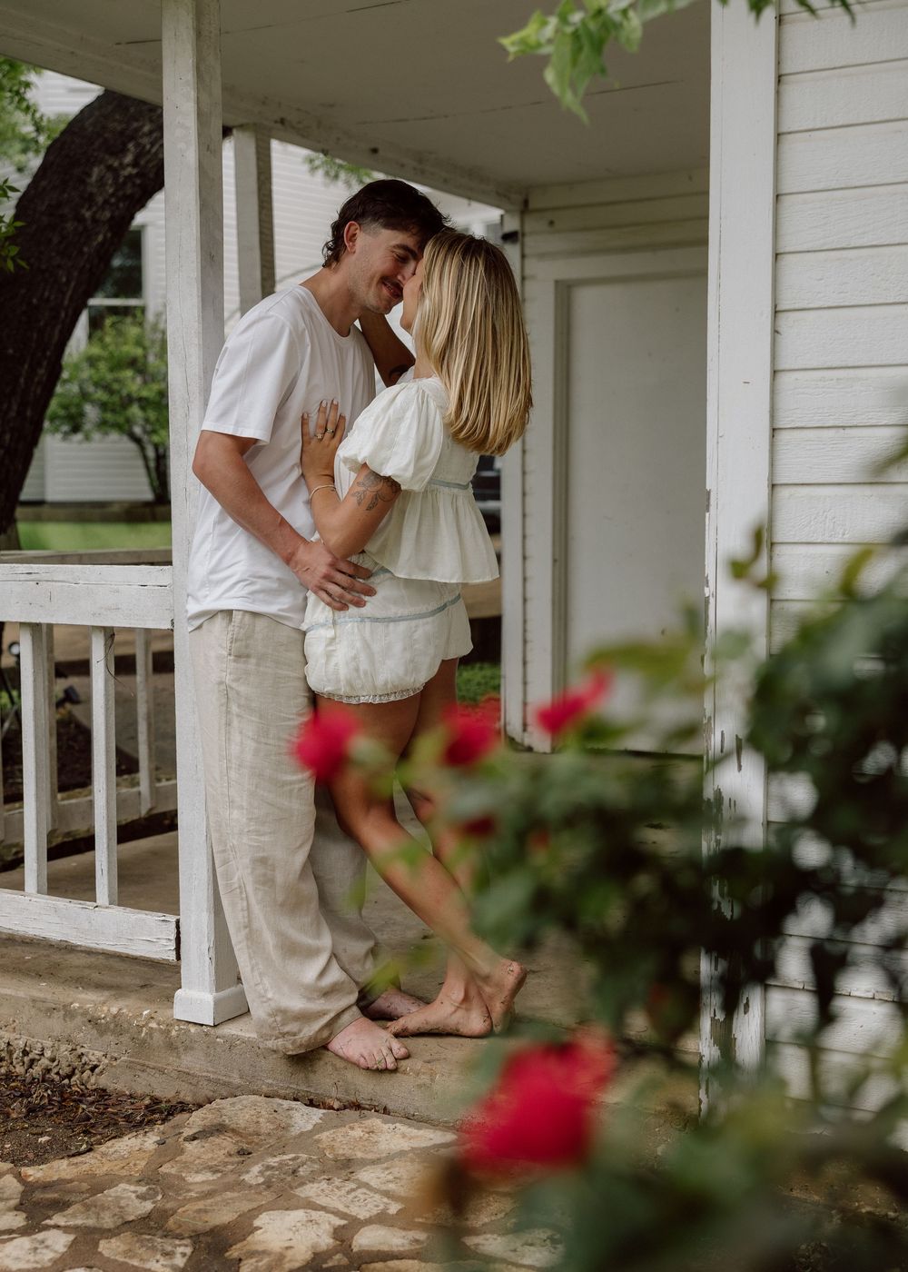 A couple shares a romantic kiss on a white porch surrounded by lush greenery and red flowers.