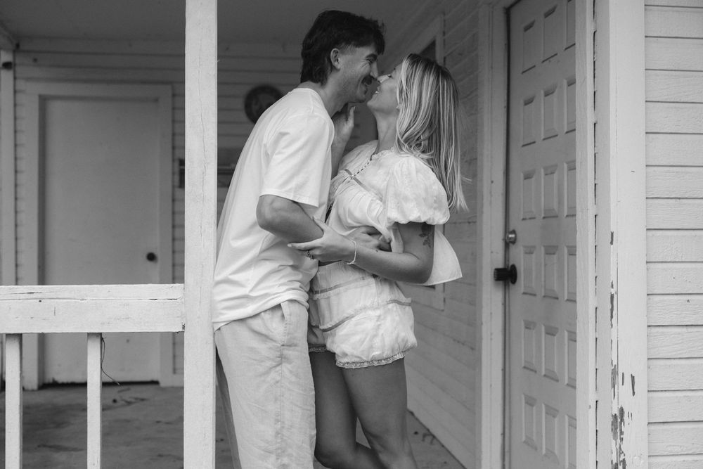 Couple sharing a romantic kiss on a porch of a white house in black and white photo.