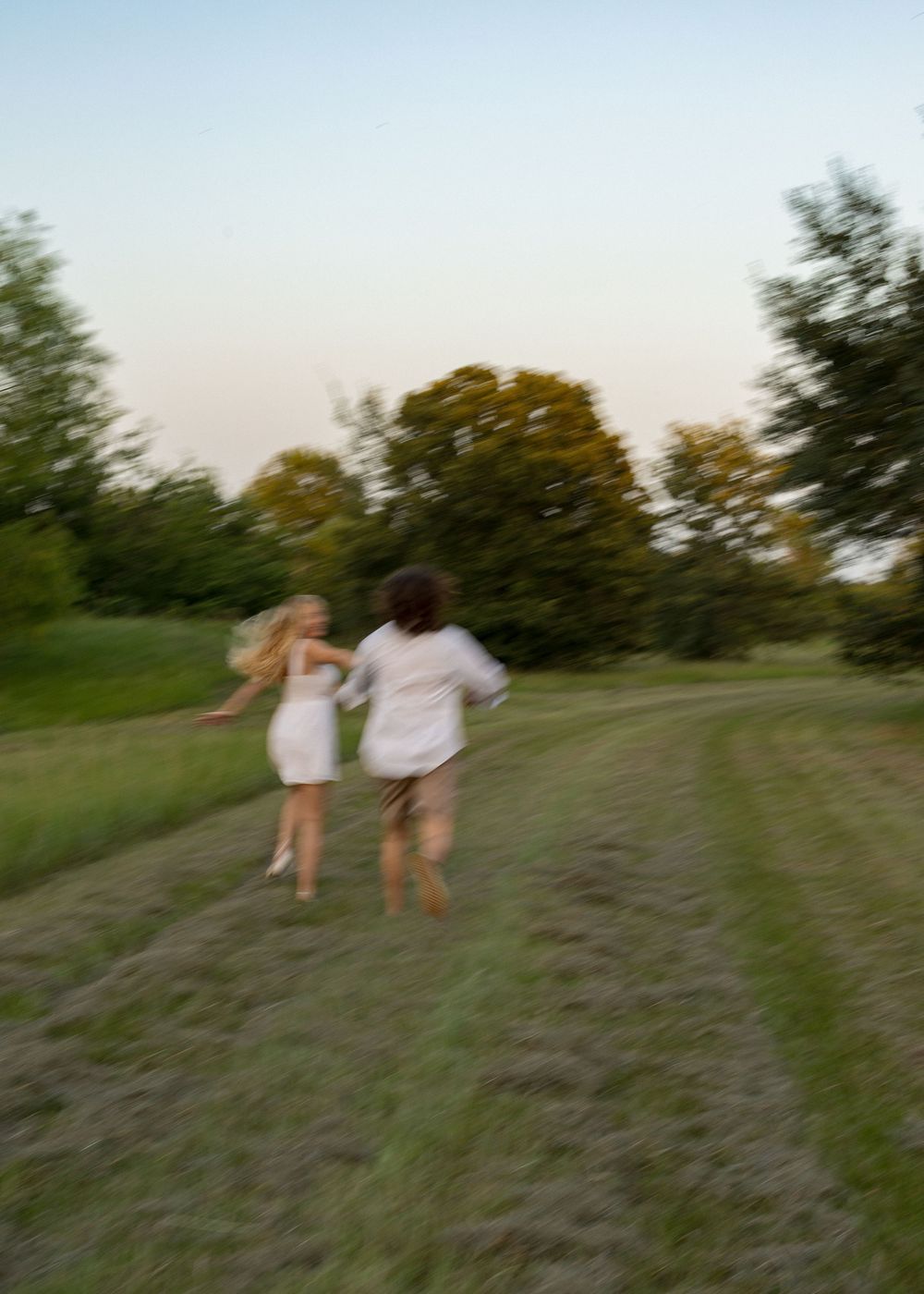 Two people in white outfits running together through a lush green field at dusk.