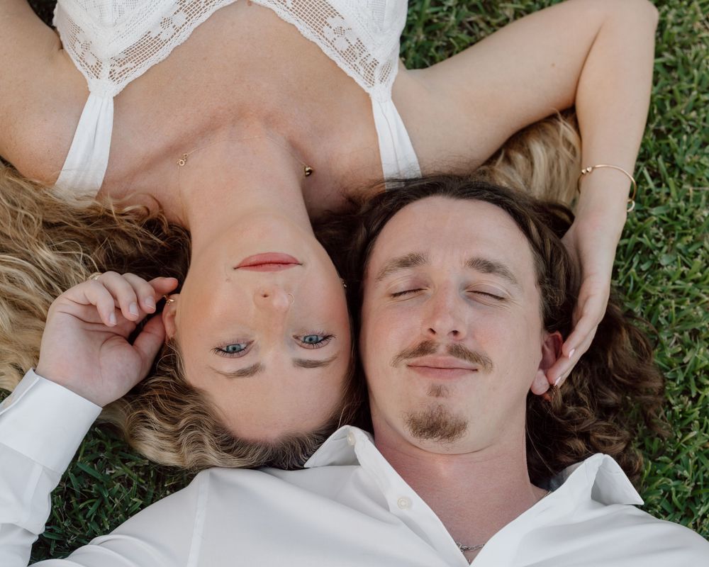 Couple lying on green grass, woman in white dress with blonde hair, man in white shirt with eyes closed, romantic outdoor photo.