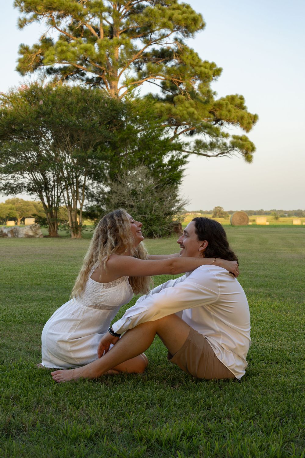 Couple sitting on green grass at sunset, smiling at each other, wearing white outfits with trees in background.