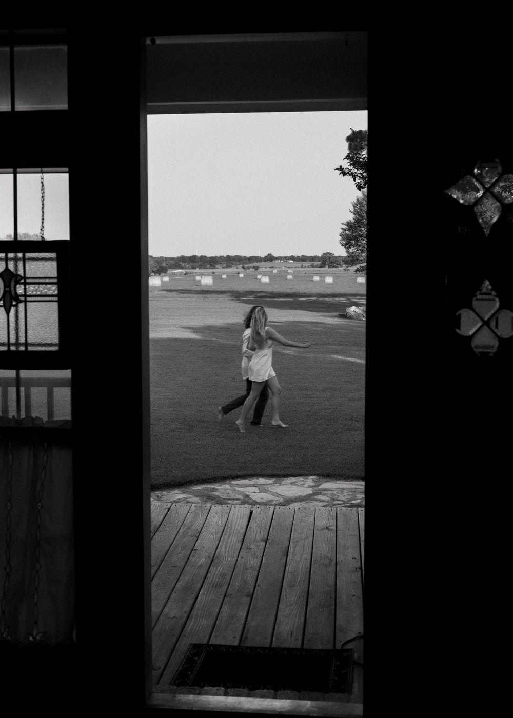 Black and white photo of a child playing outdoors, viewed through a doorway with a wooden deck and open field beyond.