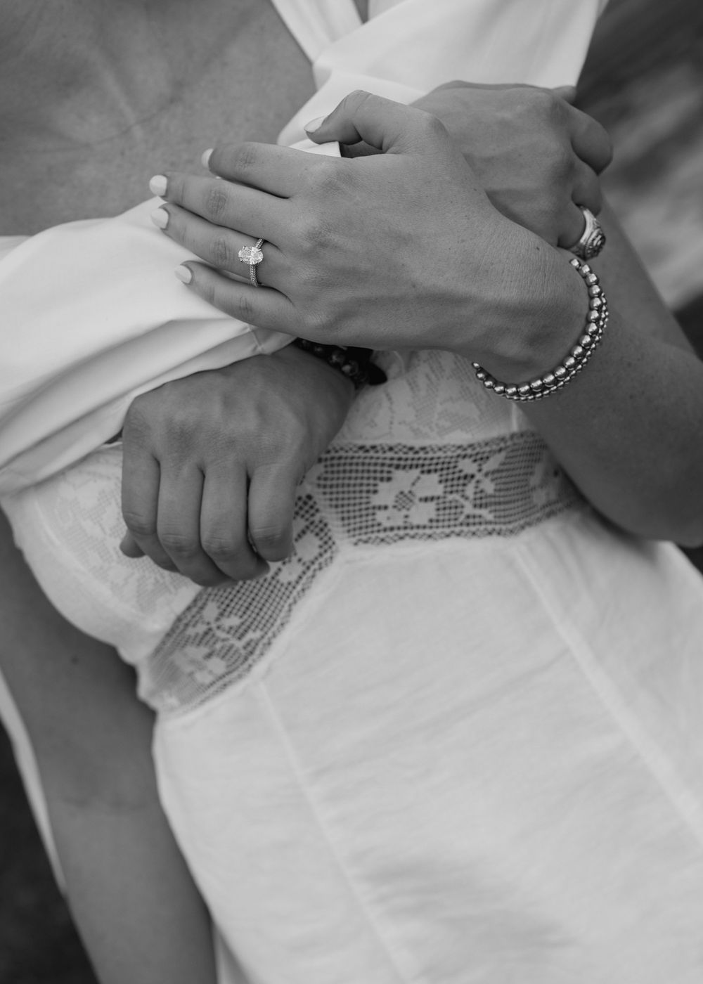 Close-up of couple holding hands, woman wearing engagement ring and man wearing bracelet, in black and white.