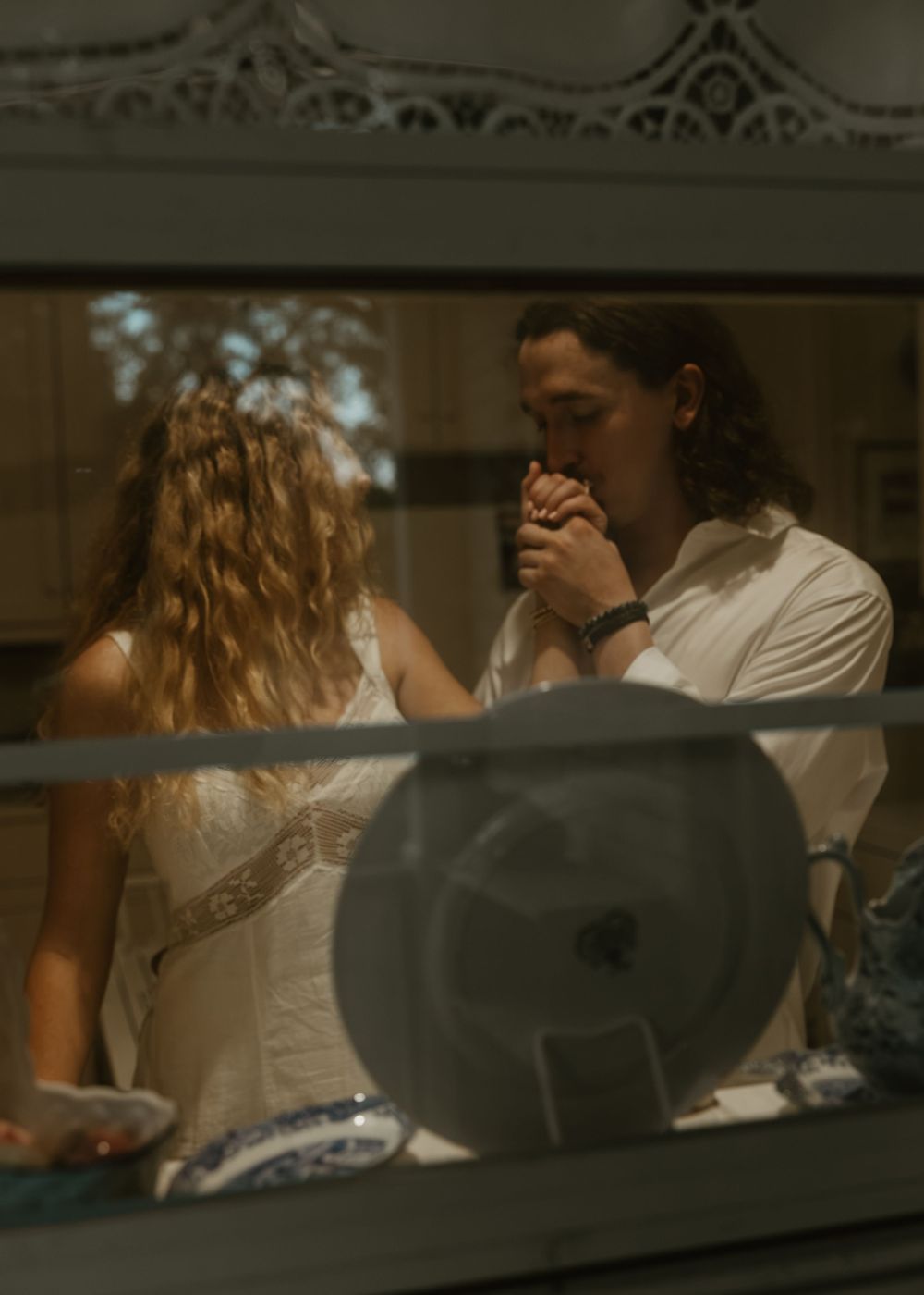 A man with long hair kisses a woman's hand tenderly, both dressed in white, viewed through a window reflection.