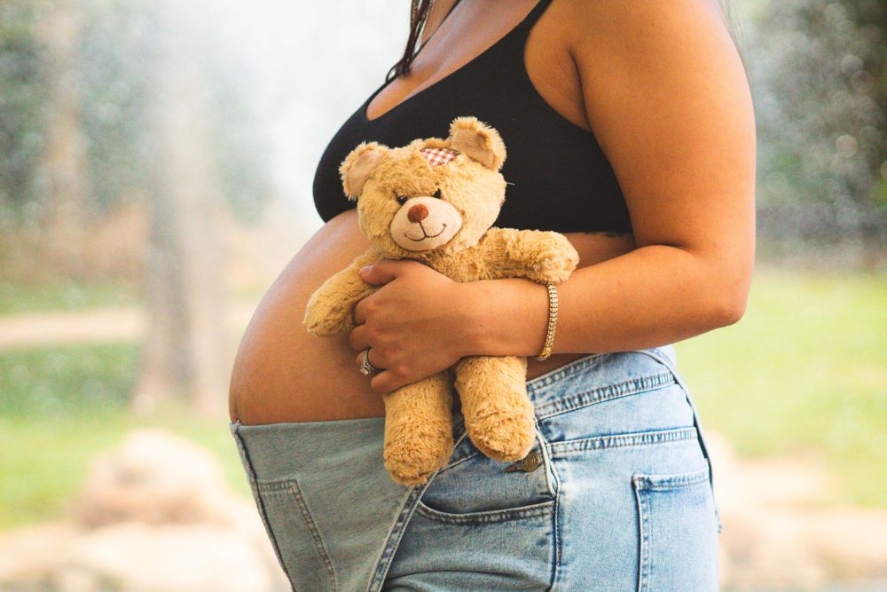 Side view of pregnant woman in black top holding teddy bear against her baby bump.