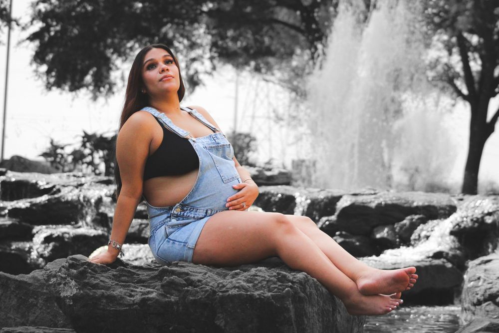 Expectant mother in overalls sitting on rocks near a fountain during maternity photoshoot.