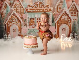 Baby's first birthday cake smash with gingerbread house backdrop and pink cake, wearing gold romper, smiling joyfully.