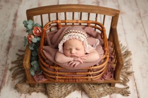 Newborn baby sleeping in rustic wooden crib with pink bonnet and floral decorations.