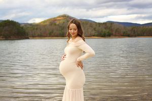 Pregnant woman in cream dress posing by a lake with mountain backdrop during maternity photoshoot.
