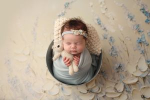 Sleeping newborn baby wrapped in blue swaddle inside a bucket, holding a stuffed animal, wearing a floral headband.