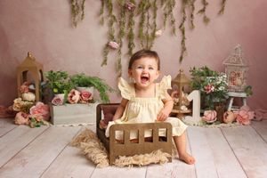 Baby girl in yellow dress sits in miniature wooden bed laughing, surrounded by flowers and greenery in a pink studio.