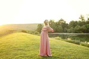 Woman in pink maxi dress standing on green grass by a pond at golden hour sunset.