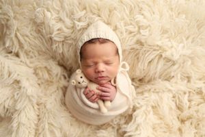 Sleeping newborn baby wrapped in cream swaddle holding knit bear toy on fluffy white rug.