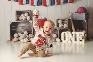 Baby boy in baseball jersey celebrates first birthday with ONE sign and baseball decorations.