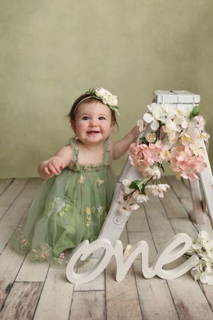 Smiling baby girl in green floral dress with flower crown beside white ladder and “one” sign for first birthday.