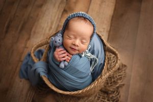 Sleeping newborn baby wrapped in blue swaddle cloth wearing knit bonnet, nestled in wicker basket with teddy bear.