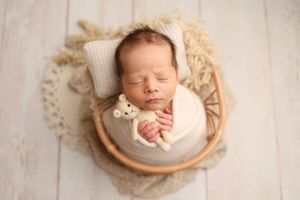 Sleeping newborn baby wrapped in cream swaddle holding tiny teddy bear in wicker basket with knit bonnet.