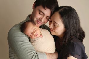 New parents smiling and holding their sleeping newborn baby wrapped in cream swaddle cloth during photo session.