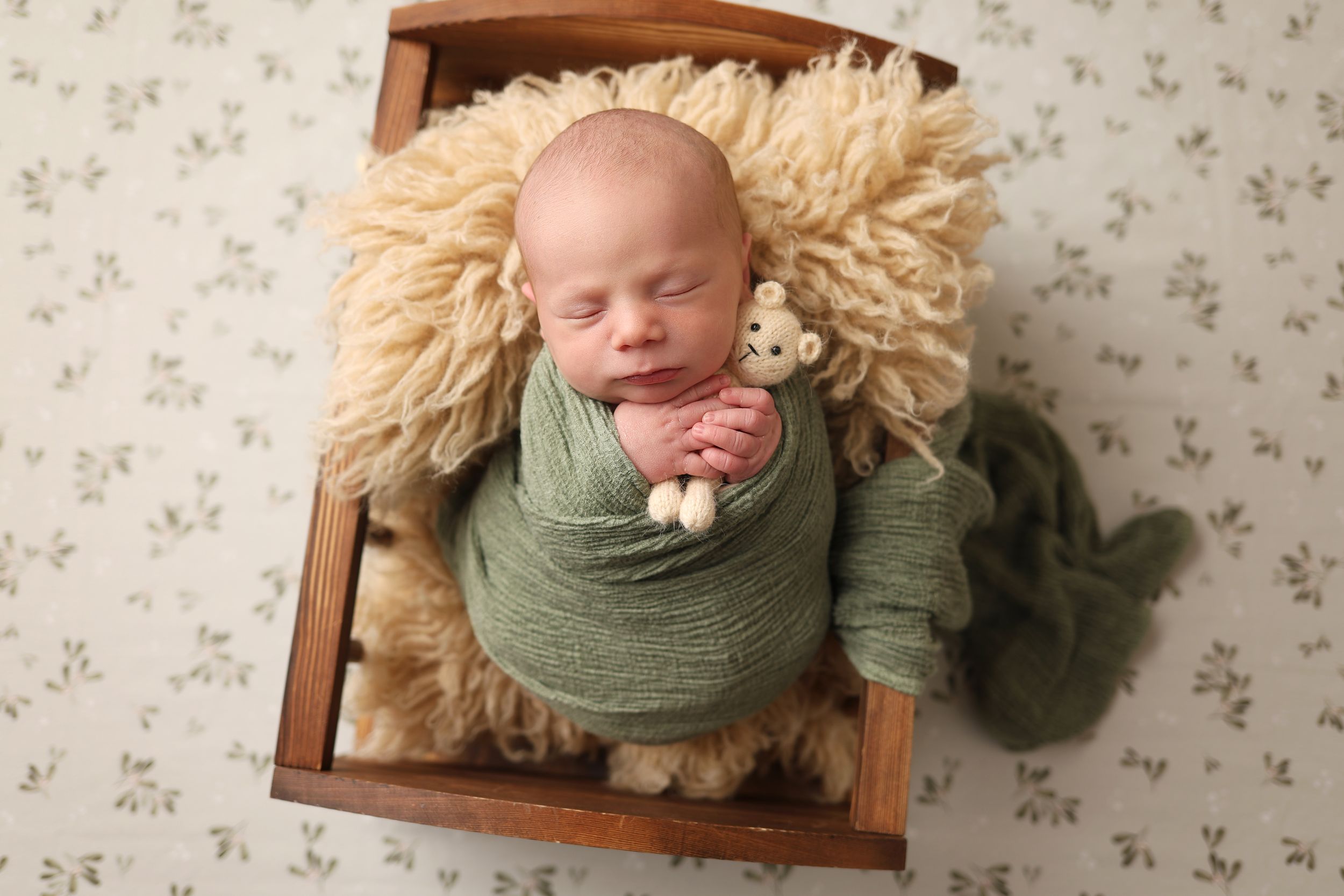 Newborn baby wrapped in green swaddle holding tiny bear, sleeping in wooden box with fluffy blanket.