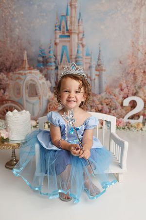 Toddler girl in blue Cinderella princess dress with tiara and wand sits on bench at Disney-themed 2nd birthday photo shoot.