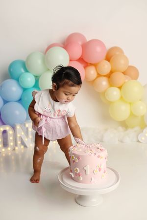 Baby girl in pink outfit smashing her first birthday cake surrounded by pastel rainbow balloon arch.