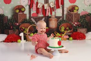 Baby in red outfit celebrating first birthday smashing a cake with apple-themed decorations.