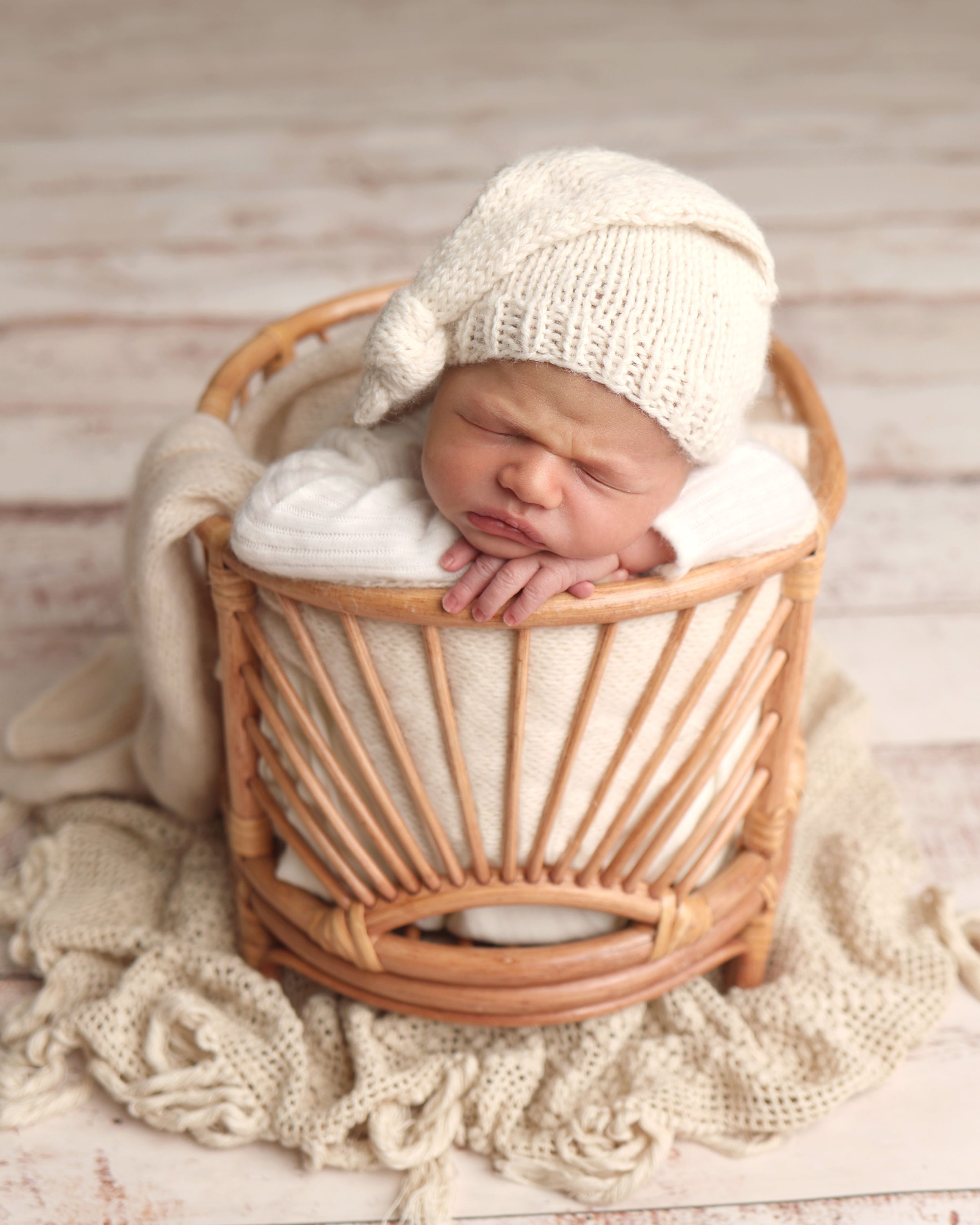 Sleeping newborn baby in cream knit hat posed in rattan basket with soft white wrap for professional newborn photography.