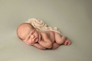 Peaceful newborn baby sleeping curled up on sage green backdrop with cream textured wrap.