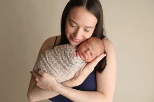 A mother tenderly holds her swaddled newborn baby close to her cheek, smiling softly against a neutral background.