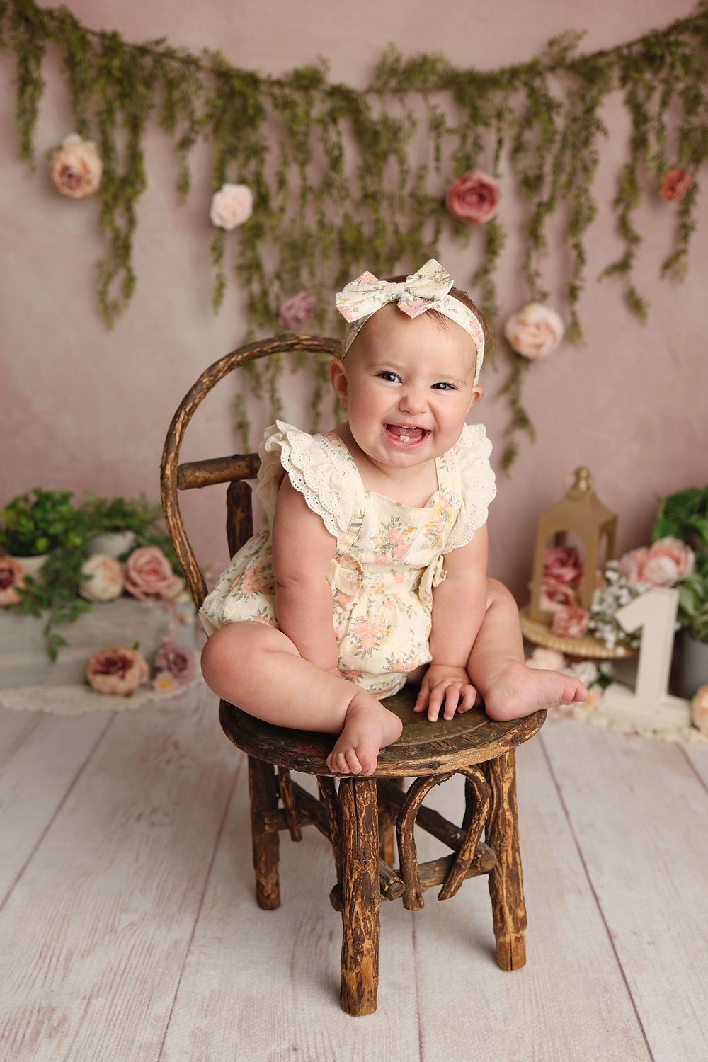 Smiling baby girl in floral romper and bow headband sits on rustic wooden chair with flower backdrop.