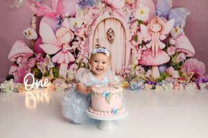 Baby girl in blue dress smiles at first birthday cake smash with fairy tale backdrop and glowing One sign.