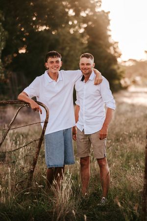 Two men in white shirts smile together in a golden-hour field, one leaning on a rusty gate.