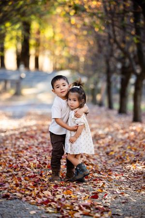 Two young siblings share a warm hug on a leaf-covered path surrounded by colorful autumn trees.